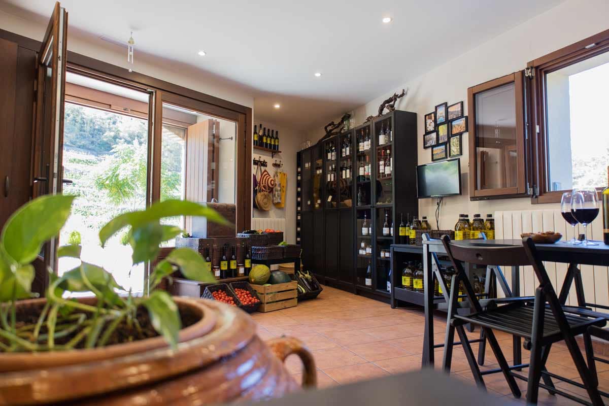 Photo of the agrobotiga, where you can see an open glass door, cabinets with glass doors and black stands with different bottles of wine, another pond with oil bottles, boxes with tomatoes, boxes with melons, boxes with aubergines...