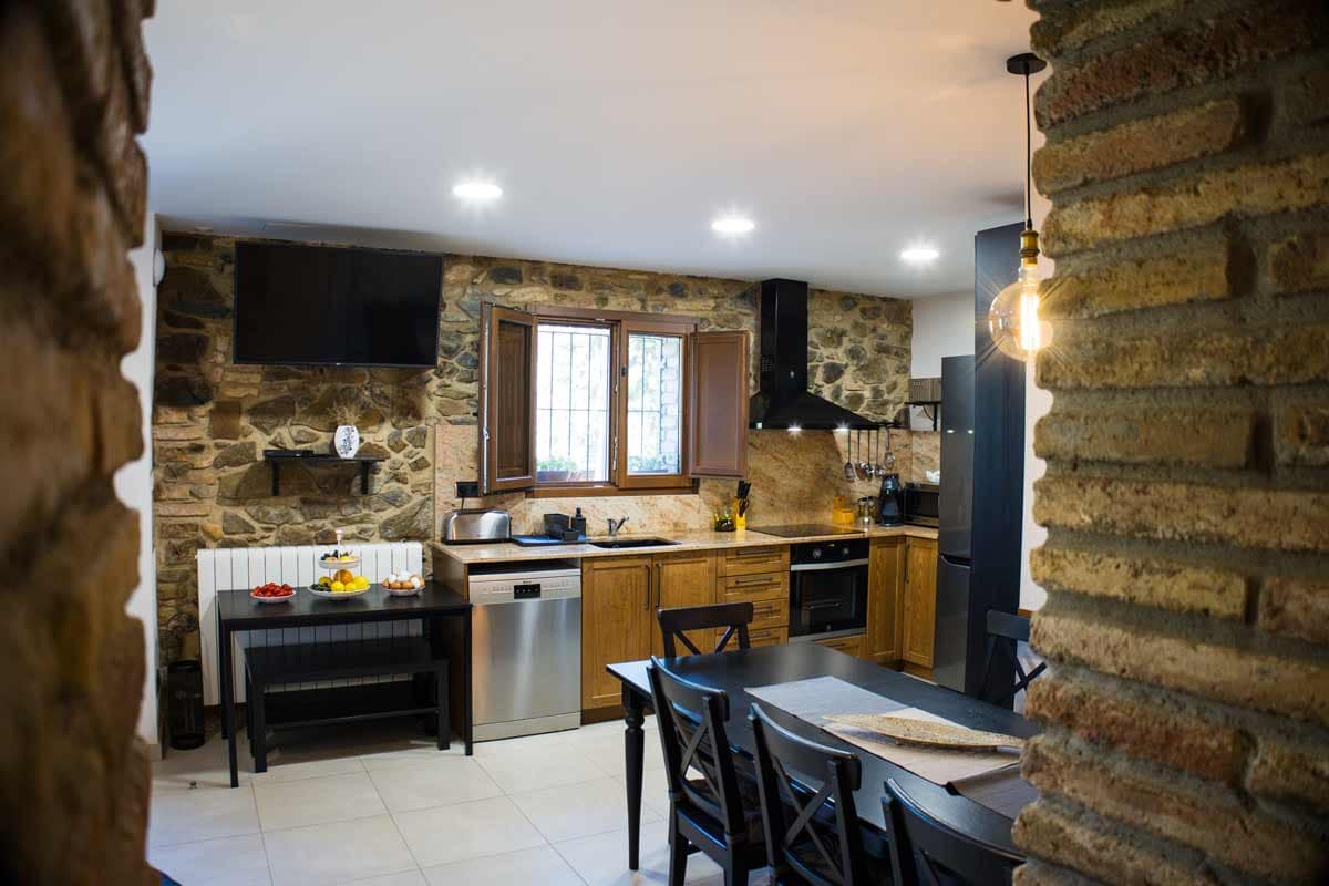 Fotografia general de la cuina on la paret és de pedra vista, on es veu una taula llarga de color negre, cadires del mateix color, una vitroceràmica, un llavaplats, una televisió, una nevera...