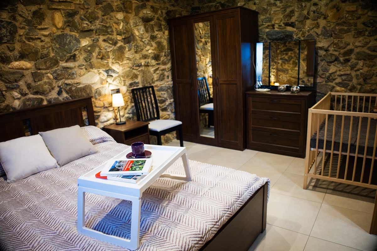 Habitació de matrimoni amb llit doble, dos coixins amb una coixinera llisa de color gris, un capçal de fusta, un armari de fusta amb un espill petit, una llum a la tauleta de nit, un bressol per infants...