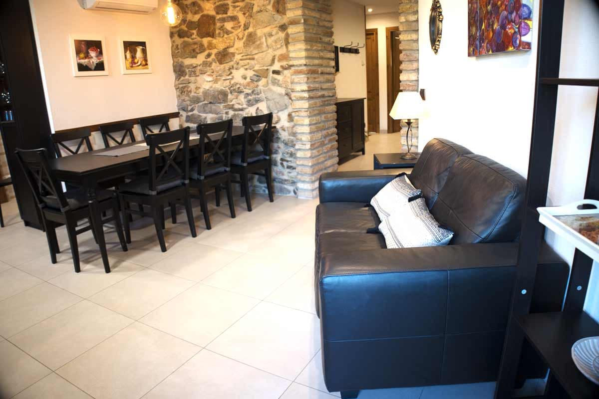 Dining - living room Sala on hi ha una taula llarga i cadires negres, un sofà de pell negra, amb coixins blancs i detalls negres, parets llises i una amb pedra vista.