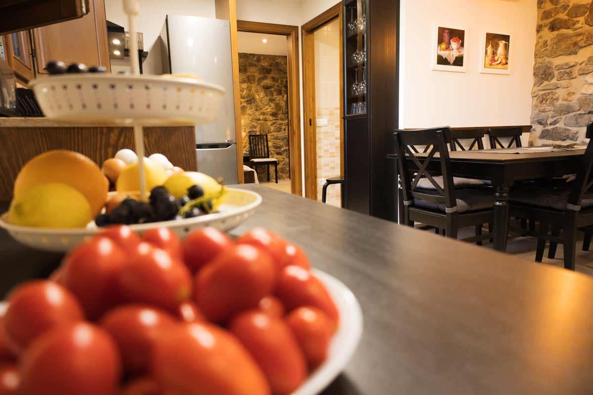 A detailed photography where fruits and vegetables are seen unfocused and you can see the dining room with black table and chairs, a white wall with handmade still life paintings hanging and a wall with exposed stone.