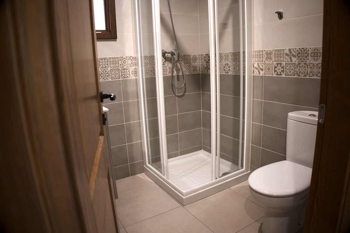 Bany on hi ha un lavabo, una dutxa tancada amb una cristallera vista, una paperera, una pica amb un moble de color fusta clar amb dos calaixos, estanteries, un espill i una tovallola color beix.