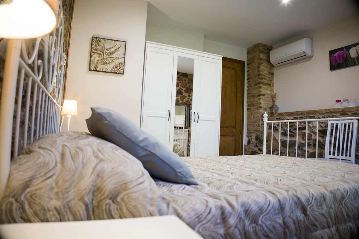 Double room Habitació de matrimoni amb llit doble, un coixi amb una coixinera llisa de color gris, un capçal de ferro pintat de color blanc, un armari blanc amb un espill petit, dues llums a les tauletes de nit i un quadre d'una fotografia on hi ha una flor de color violeta i l'altra de color blanc.