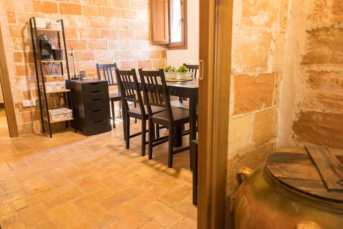 Sala petita amb el terra i les parets de totxanes vistes, on hi ha mobles de color negre, com una taula i cadires, i un armariet amb calaixos, una cafetera de color negre...