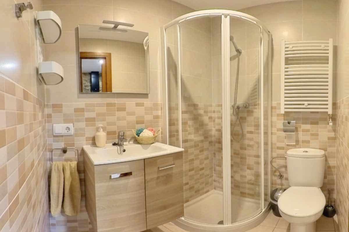 Bany de l’allotjament rural Bany on hi ha un lavabo, una dutxa tancada amb una cristallera vista, una paperera, una pica amb un moble de color fusta clar amb dos calaixos, estanteries, un espill i una tovallola color beix.