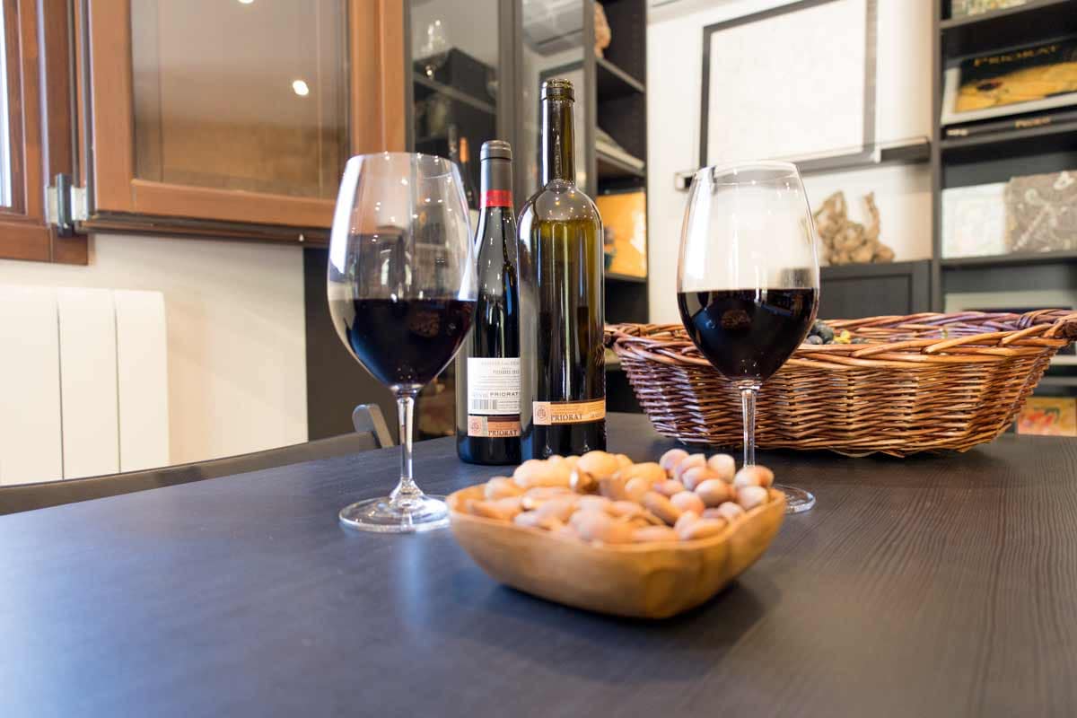 Fotografia detall de dues copes de vi amb vi negre i diferents fruits secs dintre d'un recipient de fusta, dues botelles de vi negre i una safata més enrere.