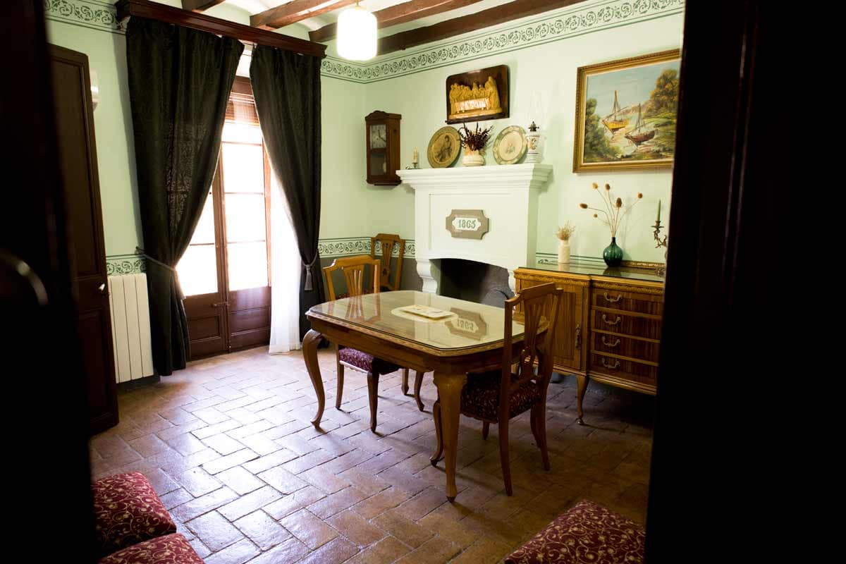 Room where you can see a fireplace, in the center there is a wooden table with a crystal above, two wooden chairs with red stamped pillows, a door with windows that enters the light of the outside, a wall clock, and other decorations on the wall such as paintings, plates and a hand painted border of dark green.