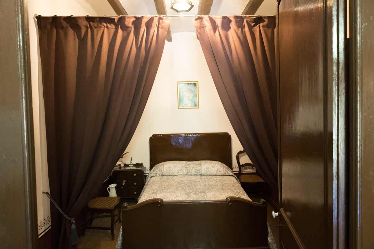 Room where there is a double bed in the center with a picture hanging in the middle of the wall, two chairs, a night table and some open curtains.